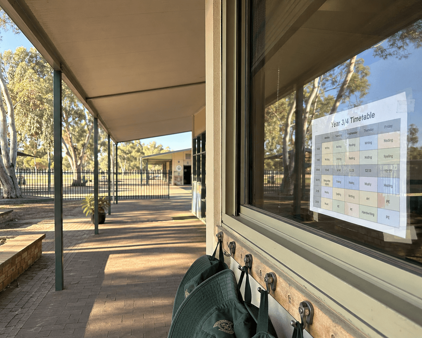 A covered walkway at an Australian primary school with gum trees, sun hats on hooks, and a Year 3/4 timetable taped inside a classroom window.