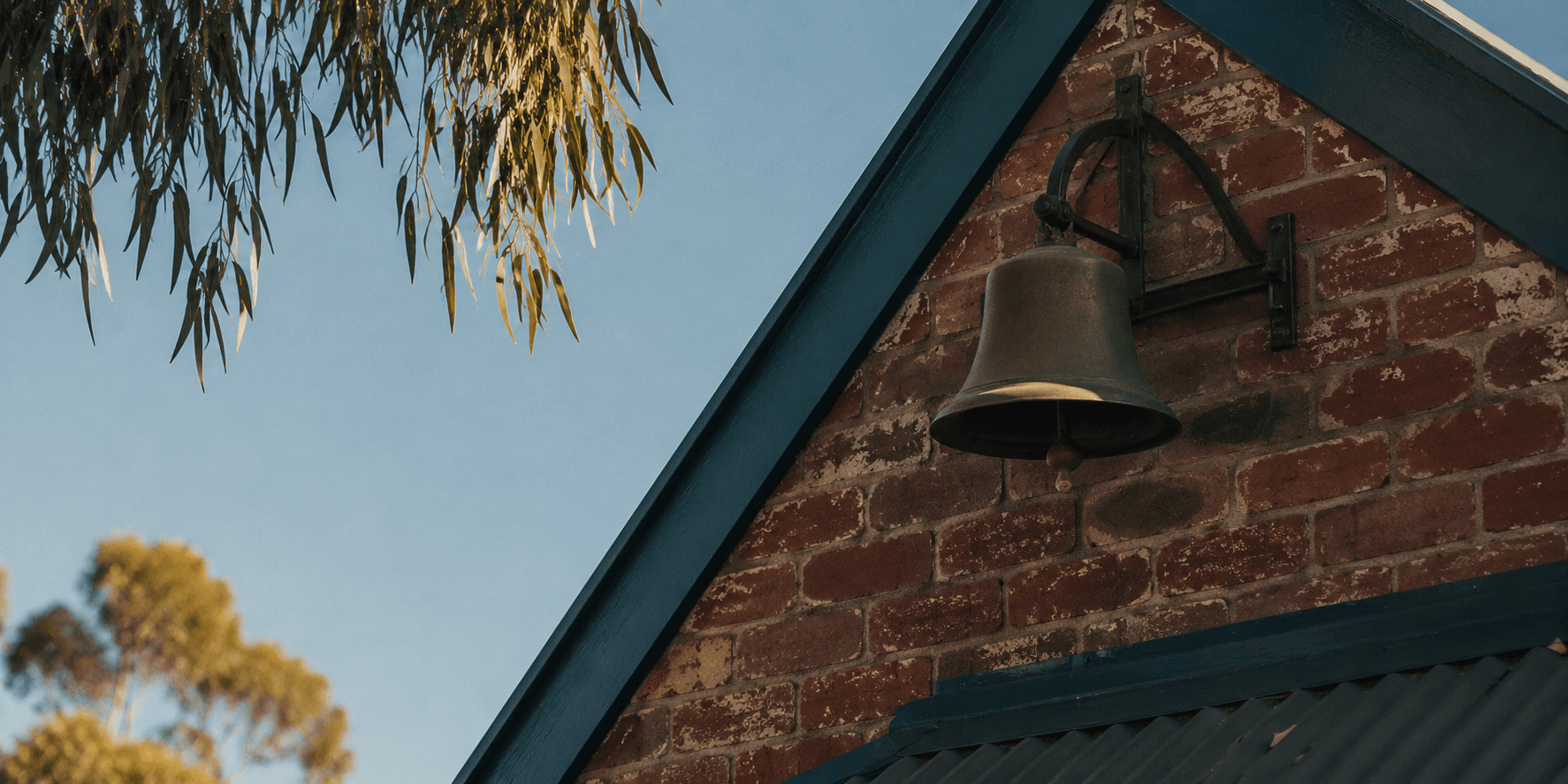 A brass school bell on a brick gable catching the first morning sun, gum leaves at the edge of frame.