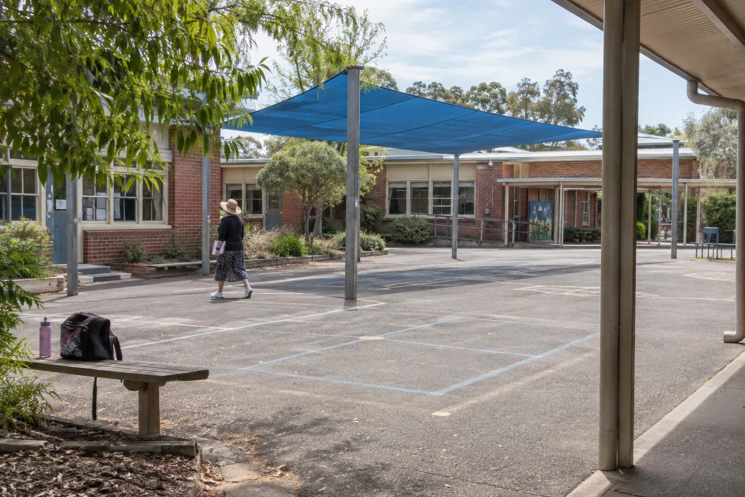 A teacher walking across a primary school courtyard beneath a blue shade sail.